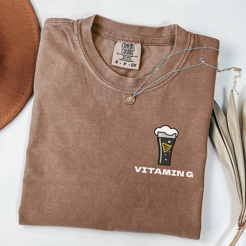 May include: A brown Comfort Colors t-shirt featuring a beer graphic and the words "VITAMIN G". A silver necklace with a pendant rests on the shirt. The t-shirt is folded on a white surface.