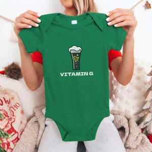 May include: A green baby bodysuit with short sleeves. The front features a graphic of a pint of beer with the text "VITAMIN G" in white. The bodysuit is made of a soft material.