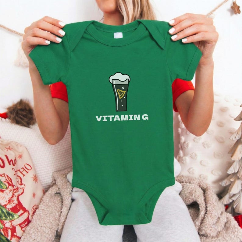 May include: A green baby bodysuit with short sleeves. The front features a graphic of a pint of beer with the text "VITAMIN G" in white. The bodysuit is made of a soft material.