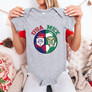 May include: A gray baby bodysuit featuring a soccer ball design split into two halves. One half displays the USA logo with stars, the other half shows the Mexican emblem. The words "USA" and "MEX" are arched above the ball.