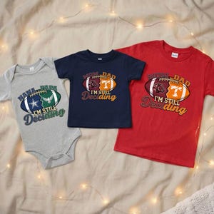 May include: Three football-themed shirts and a onesie. The gray onesie has a football graphic with the text "Nana, Papa, I'm Still Deciding." The navy blue shirt and red shirt have a football graphic with the text "Mom, Dad, I'm Still Deciding."