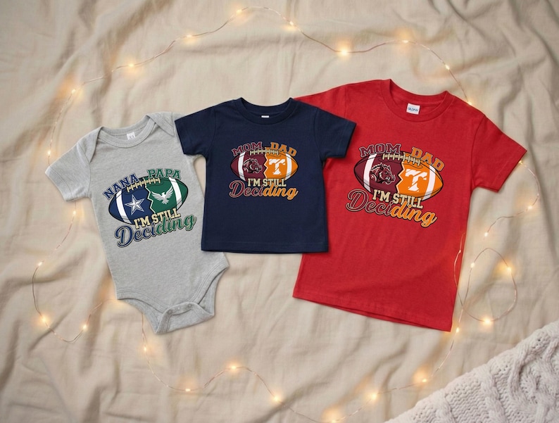 May include: Three football-themed shirts and a onesie. The gray onesie has a football graphic with the text "Nana, Papa, I'm Still Deciding." The navy blue shirt and red shirt have a football graphic with the text "Mom, Dad, I'm Still Deciding."