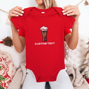 May include: A red baby bodysuit with a cartoon pint of stout beer graphic and the words "CUSTOM TEXT" in white. The beer graphic has a creamy head and a gold harp design. The bodysuit is short-sleeved.