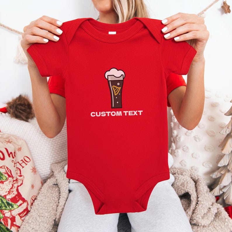 May include: A red baby bodysuit with a cartoon pint of stout beer graphic and the words "CUSTOM TEXT" in white. The beer graphic has a creamy head and a gold harp design. The bodysuit is short-sleeved.