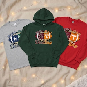 May include: Three football-themed shirts: a gray t-shirt, a green hoodie, and a red sweatshirt. Each shirt features a football graphic with text. The gray shirt says "Nana, Pa, I'm Deciding". The green hoodie says "Mom, Dad, I'm Still Deciding". The red sweatshirt says "Mom, Dad, I'm Still Deciding".