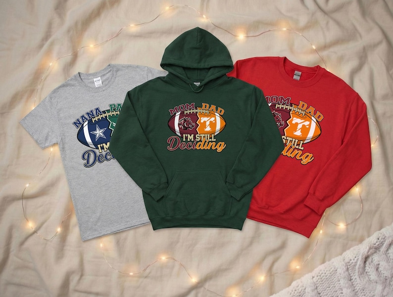 May include: Three football-themed shirts: a gray t-shirt, a green hoodie, and a red sweatshirt. Each shirt features a football graphic with text. The gray shirt says "Nana, Pa, I'm Deciding". The green hoodie says "Mom, Dad, I'm Still Deciding". The red sweatshirt says "Mom, Dad, I'm Still Deciding".
