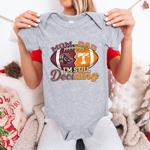 May include: A gray baby onesie with a football-themed graphic. The graphic includes the words "MOM DAD" above a football design with a Razorback and a "T" logo. Below the football is the text "I'M STILL Deciding".