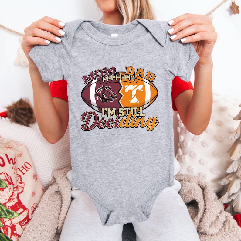 May include: A gray baby onesie with a football-themed graphic. The graphic includes the words "MOM DAD" above a football design with a Razorback and a "T" logo. Below the football is the text "I'M STILL Deciding".