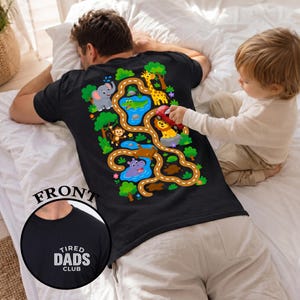 May include: Black t-shirt with a colorful jungle-themed design on the back, featuring animals and a road. The front of the shirt has the text "TIRED DADS CLUB" in white. A child plays with a toy car on the shirt.