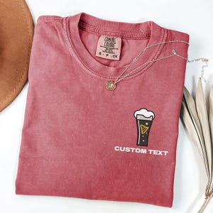 May include: A faded red Comfort Colors t-shirt featuring a beer graphic and the words "CUSTOM TEXT". A silver necklace with a pendant is draped across the shirt.