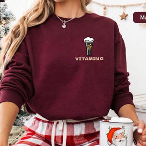 Puede incluir: Sudadera granate con un peque&ntilde;o gr&aacute;fico de una pinta de cerveza negra y la palabra "VITAMIN G" impresa debajo. La sudadera es de color granate liso y tiene mangas largas. La modelo lleva un pantal&oacute;n de pijama a cuadros rojos y blancos.