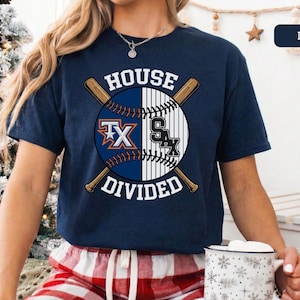 May include: Navy blue t-shirt featuring a baseball design with the text "HOUSE DIVIDED." The design includes crossed baseball bats and a baseball split in half, with the logos of two baseball teams. The shirt is short-sleeved.