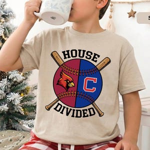 May include: A tan t-shirt featuring a baseball design with the text "HOUSE DIVIDED." The baseball is split into two sections, one maroon with a cardinal bird and the other blue with a white "C." Two crossed baseball bats are behind the baseball.