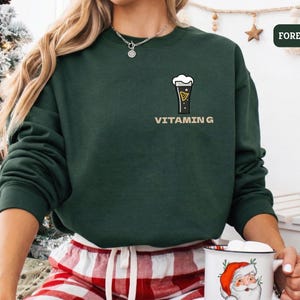 Puede incluir: Una sudadera verde bosque con un gr&aacute;fico de un vaso de cerveza y el texto "VITAMIN G". La sudadera es de color liso y tiene mangas largas. La persona lleva un pantal&oacute;n de pijama a cuadros rojos y blancos y sostiene una taza de Pap&aacute; Noel.