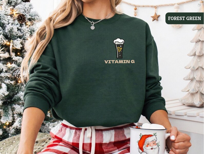 Puede incluir: Una sudadera verde bosque con un gr&aacute;fico de un vaso de cerveza y el texto "VITAMIN G". La sudadera es de color liso y tiene mangas largas. La persona lleva un pantal&oacute;n de pijama a cuadros rojos y blancos y sostiene una taza de Pap&aacute; Noel.