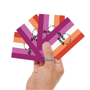May include: A set of four rectangular playing cards featuring a vertical striped pattern in shades of dark pink, light pink, white, and orange. Each card displays the black text dyke deck and the circular text she's got a good hand.