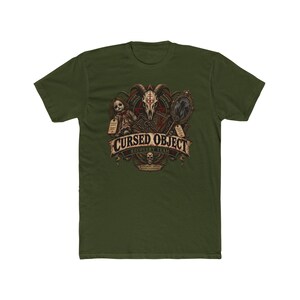 May include: An olive green short-sleeve t-shirt featuring a graphic print of a goat skull, a doll, a book, and a mirror. The text reads CURSED OBJECT RECOVERY TEAM, with additional small text surrounding the central illustration of occult items.