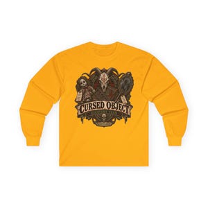 May include: A mustard yellow long-sleeve shirt featuring a graphic print of a goat skull, a doll, and a mirror. The text reads CURSED OBJECT RECOVERY TEAM, with additional small text elements integrated into the dark, detailed illustration.