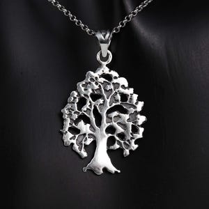 May include: A silver-colored pendant necklace featuring a detailed tree of life design. The pendant is suspended from a delicate chain, showcasing the intricate branches and leaves of the tree. The necklace is displayed against a dark background.