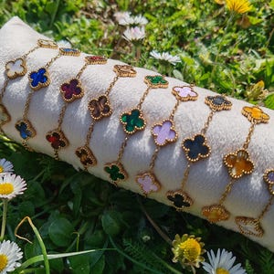 Op de afbeelding: Gouden armbanden met klavervormige hangers in diverse kleuren, waaronder wit, blauw, rood, groen en geel. De armbanden worden gepresenteerd op een wit kussen tegen een achtergrond van groen gras en bloemen.