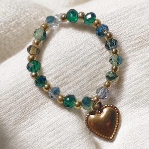 May include: A beaded ring featuring a mix of green, blue, and clear faceted beads, interspersed with small gold-colored beads. A gold-colored heart charm dangles from the ring. The ring is set against a white fabric background.