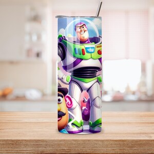 May include: A cylindrical stainless steel tumbler featuring a full-wrap graphic print of the character Buzz Lightyear in a white, green, and purple space suit, alongside a partial view of Mr. Potato Head. The tumbler includes a metal straw.