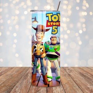 May include: A cylindrical stainless steel tumbler featuring a colorful graphic print of the characters Woody and Buzz Lightyear from the Toy Story franchise, with the text Toy Story 5 prominently displayed at the top and a metal straw inserted.