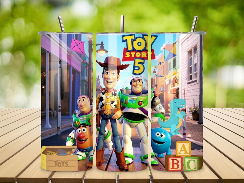May include: A cylindrical metal tumbler featuring a colorful Toy Story 5 graphic print. The design displays characters including Woody, Buzz Lightyear, Rex, and Mr. Potato Head set against a street scene with a toy box and alphabet blocks.