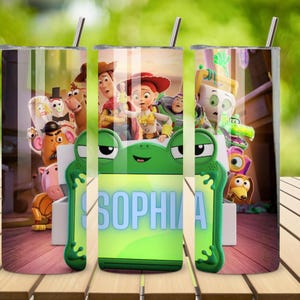Op de afbeelding: Een cilindervormige roestvrijstalen beker met een rondlopende afbeelding van Toy Story-personages, waaronder Woody, Jessie en Buzz Lightyear. Het ontwerp bevat een opvallend groen kikker-vormig kader met de naam SOPHIA in het blauw.