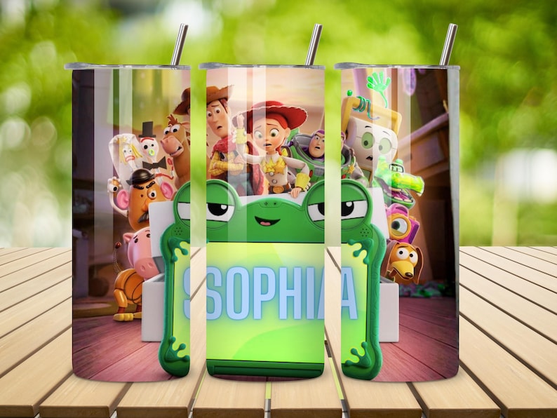 Op de afbeelding: Een cilindervormige roestvrijstalen beker met een rondlopende afbeelding van Toy Story-personages, waaronder Woody, Jessie en Buzz Lightyear. Het ontwerp bevat een opvallend groen kikker-vormig kader met de naam SOPHIA in het blauw.