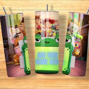 Op de afbeelding: Een cilindervormige metalen beker met een rondom lopende afbeelding van Toy Story-personages, waaronder Woody, Jessie en Buzz Lightyear. Het ontwerp bevat een groen kikker-vormig kader met de tekst ADD YOUR NAME HERE in lichtblauwe letters.