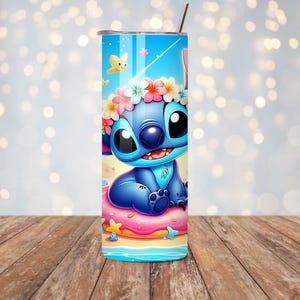 May include: A blue tumbler featuring a cartoon character with large black eyes, a flower crown, and a smiling expression. The tumbler has a blue background with butterflies and a beach scene. A silver straw is inserted into the lid.