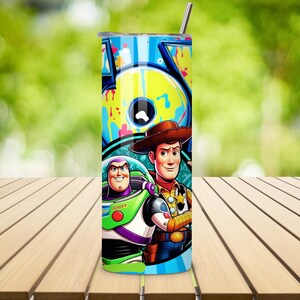 K&ouml;nnte beinhalten: Ein hoher, zylindrischer Becher mit einem Edelstahlstrohhalm. Der Becher zeigt eine farbenfrohe Grafik von Buzz Lightyear und Woody aus Toy Story, vor einem Hintergrund im Graffiti-Stil. Das Design enth&auml;lt das Wort "TOY" in gro&szlig;en Buchstaben.