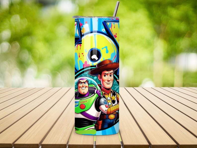 K&ouml;nnte beinhalten: Ein hoher, zylindrischer Becher mit einem Edelstahlstrohhalm. Der Becher zeigt eine farbenfrohe Grafik von Buzz Lightyear und Woody aus Toy Story, vor einem Hintergrund im Graffiti-Stil. Das Design enth&auml;lt das Wort "TOY" in gro&szlig;en Buchstaben.