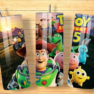 May include: A cylindrical tumbler with a metal straw featuring a Toy Story 5 graphic print. The design displays characters including Woody, Buzz Lightyear, Rex, Hamm, and Ducky against a sunset landscape. The text Toy Story 5 is visible at the top.