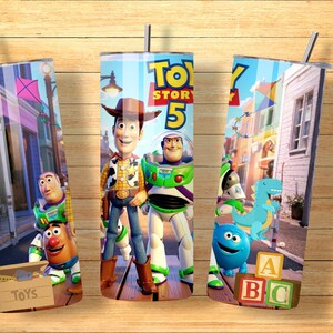May include: A cylindrical tumbler featuring a Toy Story 5 graphic with Woody, Buzz Lightyear, Rex, and Mr. Potato Head. The design includes a toy box labeled TOYS, alphabet blocks, and a street scene background. A metal straw is inserted at the top.