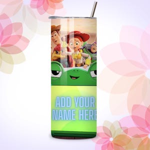 Op de afbeelding: Een cilindervormige metalen beker met rietje, voorzien van een Toy Story-personageafbeelding aan de bovenkant, een groen kikkergezicht in het midden en de tekst ADD YOUR NAME HERE in lichtblauwe letters op een limoengroene achtergrond.