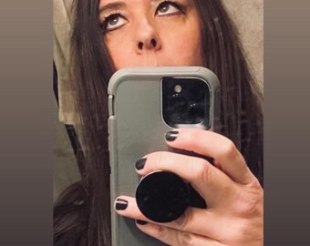 May include: A person holding a gray phone with a black pop socket. The person has dark hair and is looking upwards. The phone case is gray and the nails are painted black.