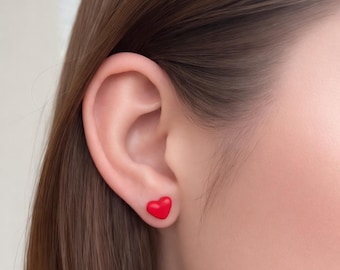 May include: A close-up of a person's ear with a small, red heart-shaped stud earring. The earring is a vibrant, solid red color. The skin tone is fair, and the hair is a medium brown.