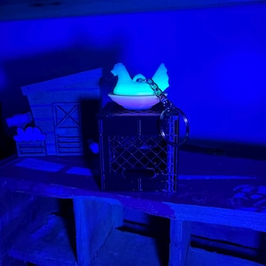 Puede incluir: Una figura de gallina blanca brillante sobre una peque&ntilde;a jaula oscura, sujeta a un llavero. La gallina est&aacute; posada en un cuenco blanco. Una granja de madera en miniatura est&aacute; en el fondo. La escena est&aacute; iluminada con luz azul.