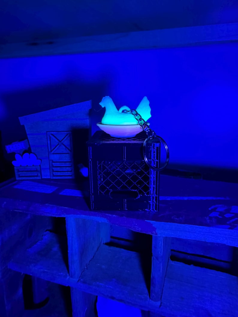 Puede incluir: Una figura de gallina blanca brillante sobre una peque&ntilde;a jaula oscura, sujeta a un llavero. La gallina est&aacute; posada en un cuenco blanco. Una granja de madera en miniatura est&aacute; en el fondo. La escena est&aacute; iluminada con luz azul.