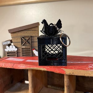Puede incluir: Un llavero negro impreso en 3D con una gallina posada sobre una caja en miniatura. La caja tiene un dise&ntilde;o de celos&iacute;a. El llavero est&aacute; sobre una superficie roja, con una estructura de madera en el fondo. Un letrero dice "FRESH".