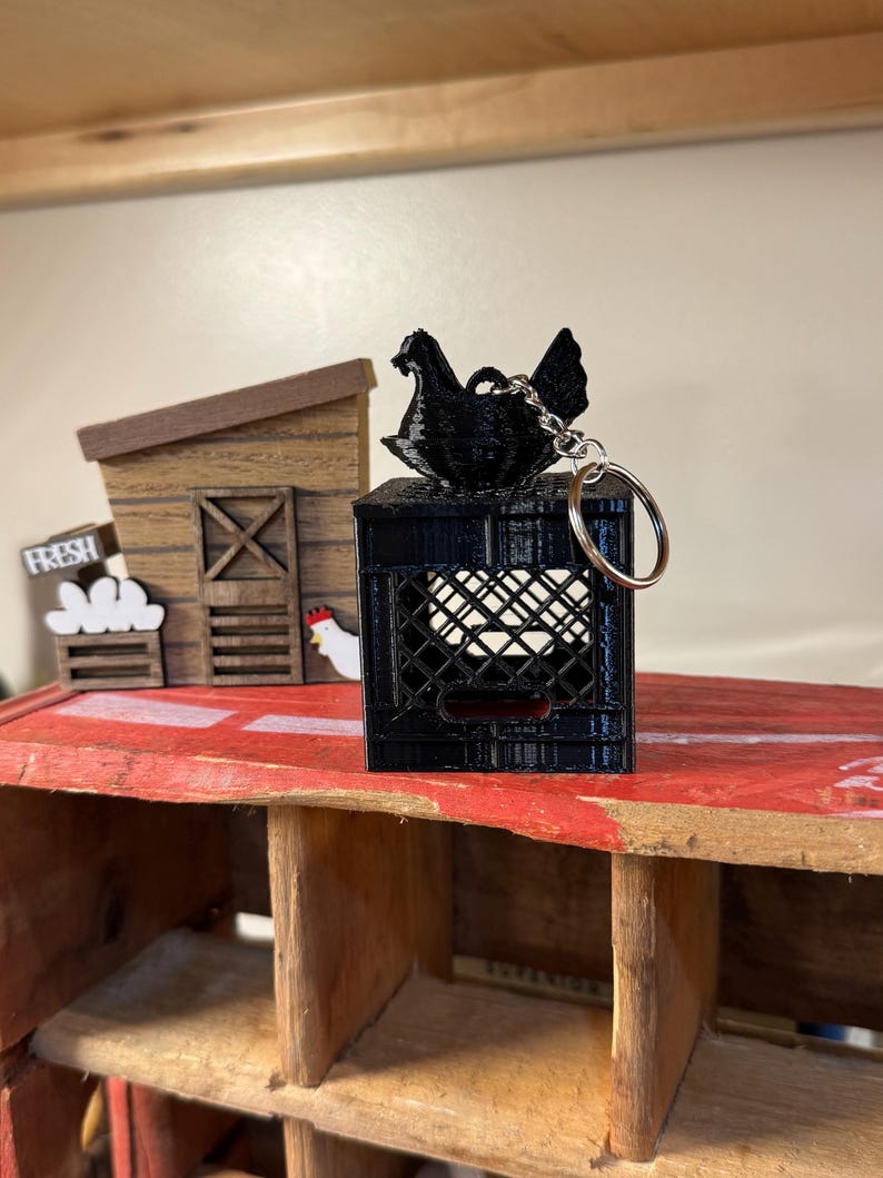 Puede incluir: Un llavero negro impreso en 3D con una gallina posada sobre una caja en miniatura. La caja tiene un dise&ntilde;o de celos&iacute;a. El llavero est&aacute; sobre una superficie roja, con una estructura de madera en el fondo. Un letrero dice "FRESH".