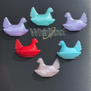 May include: Six colorful chicken-shaped magnets in shades of purple, blue, and red. Each magnet features a hen sitting in a nest-like dish. The magnets are arranged on a dark gray surface, with the Whirlpool logo visible.
