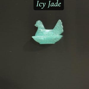 May include: A teal-colored, translucent hen figurine sitting in a nest. The text "Icy Jade" is displayed in a black rectangle above the figurine. The background is a dark gray.