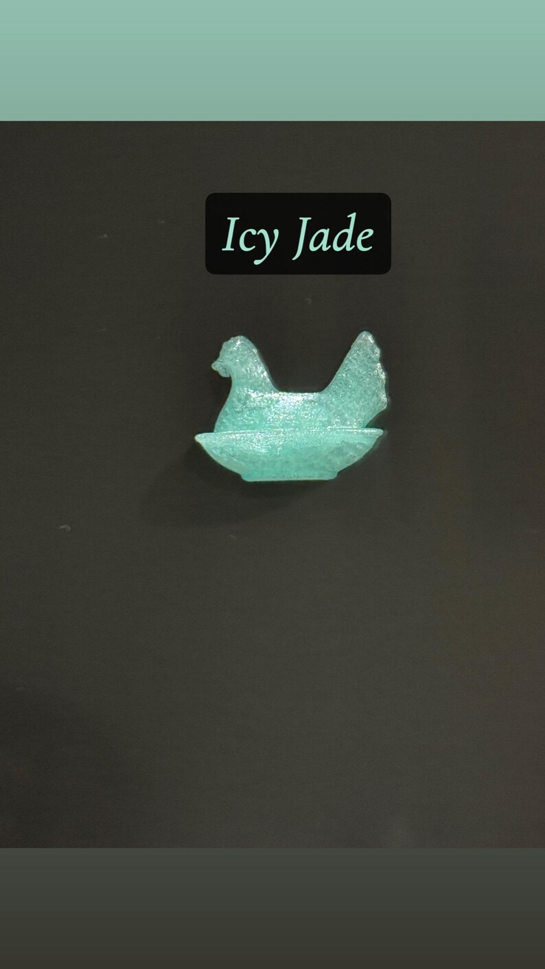 May include: A teal-colored, translucent hen figurine sitting in a nest. The text "Icy Jade" is displayed in a black rectangle above the figurine. The background is a dark gray.