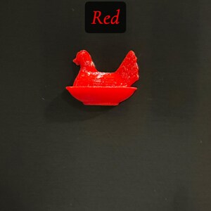May include: A bright red hen figurine sitting in a red bowl. The word "Red" is displayed in a black box above the figurine. The background is a solid black.