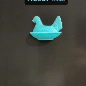 May include: A turquoise-colored hen figurine in a nest. The text "Feather Blue" is displayed in a black rectangle above the figurine. The hen is sitting in a shallow dish, with its head and tail feathers visible.
