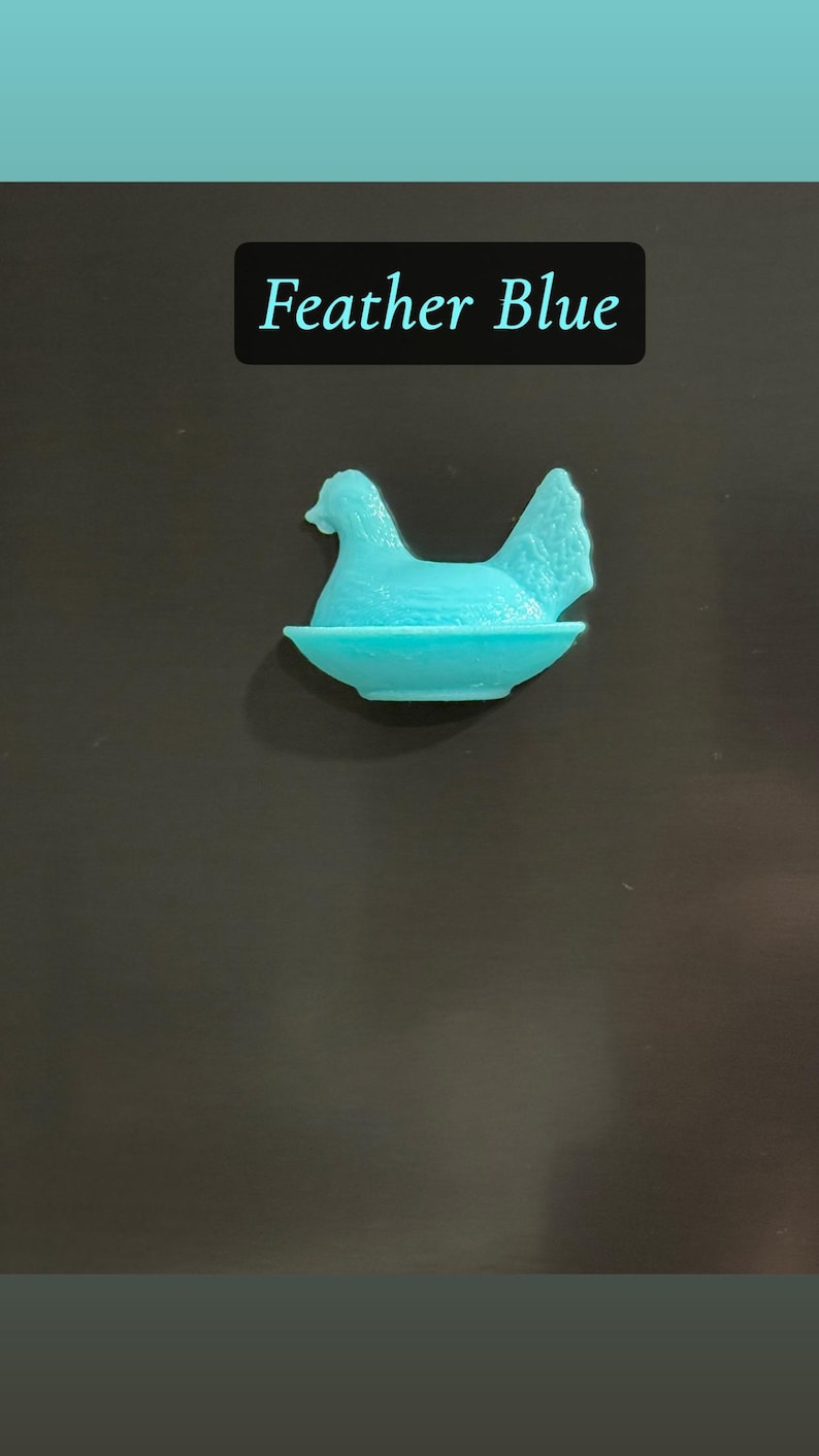 May include: A turquoise-colored hen figurine in a nest. The text "Feather Blue" is displayed in a black rectangle above the figurine. The hen is sitting in a shallow dish, with its head and tail feathers visible.