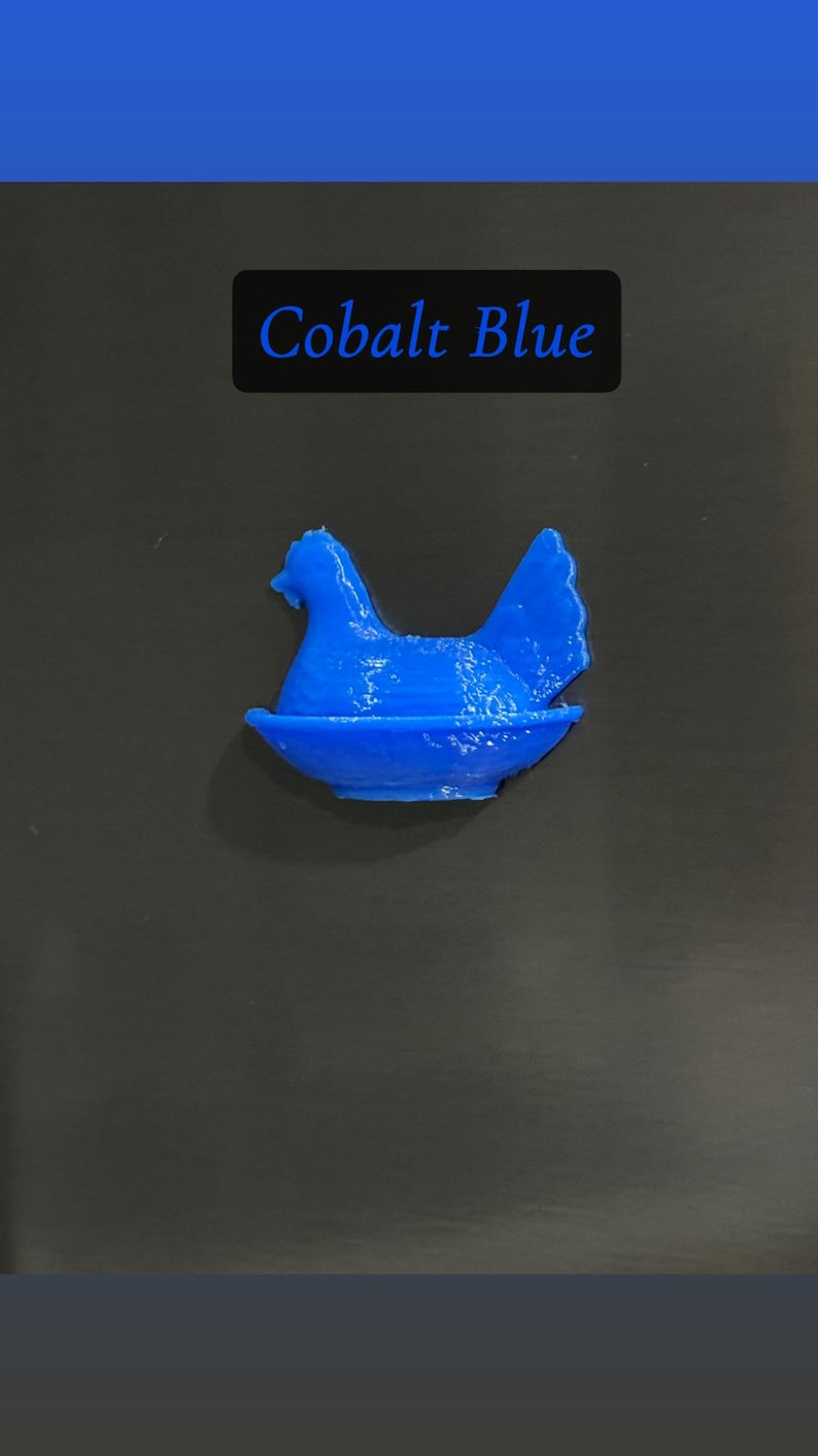 May include: A cobalt blue hen figurine in a nest. The text "Cobalt Blue" is displayed in a black rectangle above the figurine. The hen is a solid color and appears to be 3D printed.